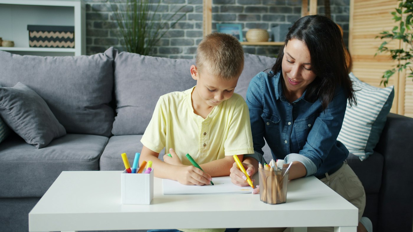 A psychologist sits beside a school-age boy as he works on a drawing task at a table