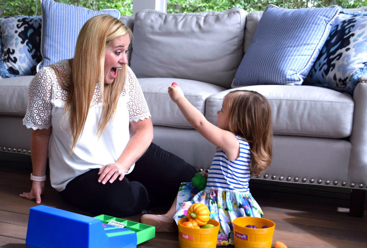A clinician sits on the floor by a grey sofa, playing with a toddler girl