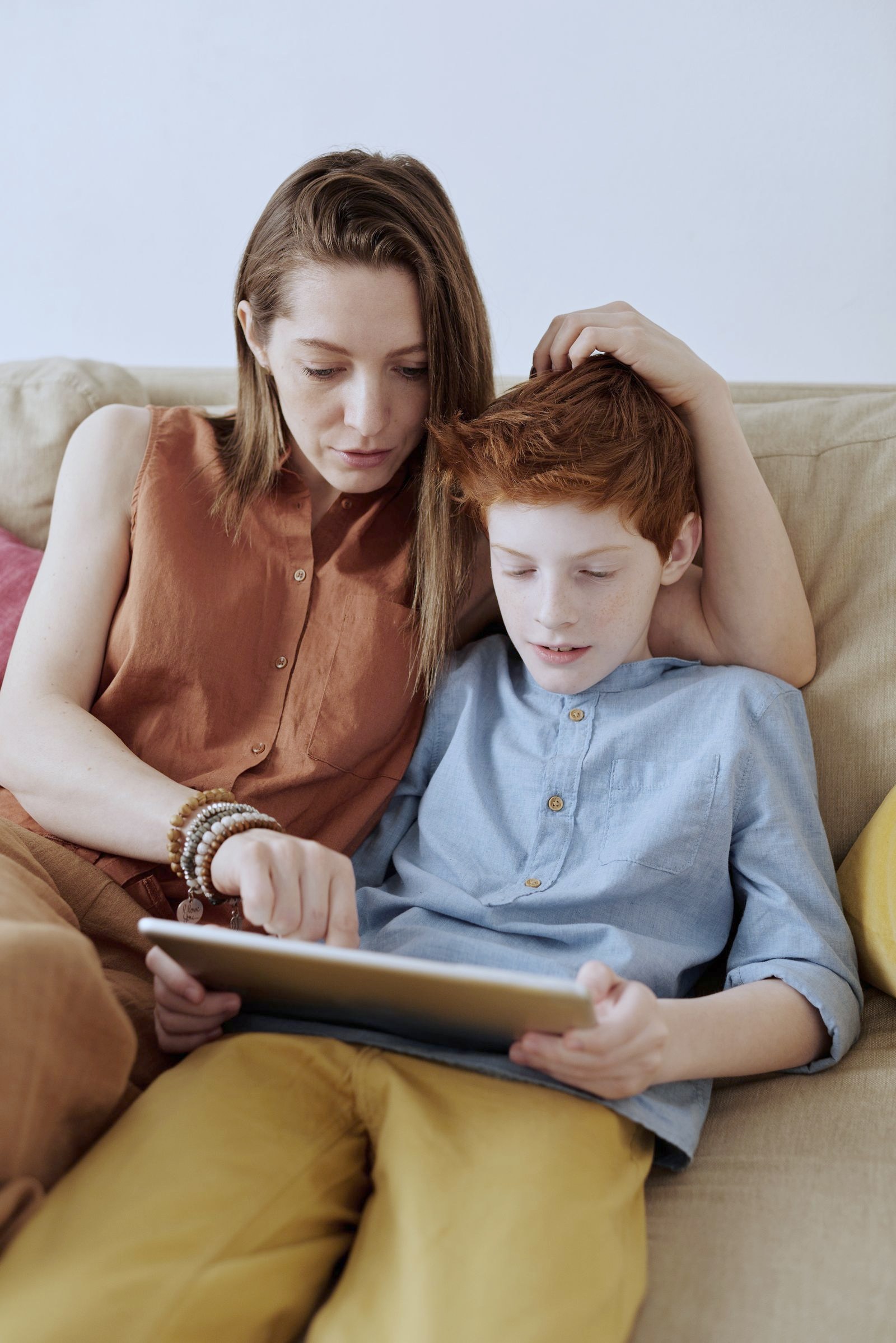 A mother and her son sitting close together on the couch, looking at a tablet